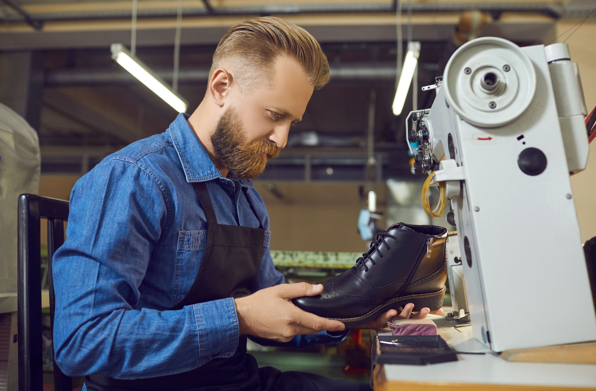 Male shoemaker sewing and checking boot on defect at footwear factory workshop